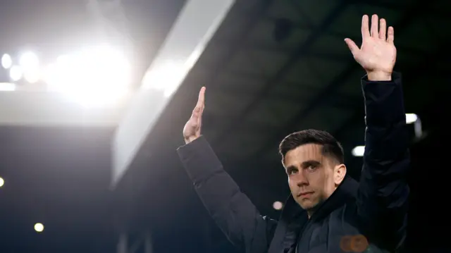 A photo of Eric Ramsay, wearing a black puffer jacket, holding his hands up in the air while the stadium lights shine in the background