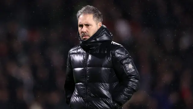 Marti Cifuentes watching his Leicester City side face Wrexham, standing in the rain wearing a shiny, black puffer jacket.