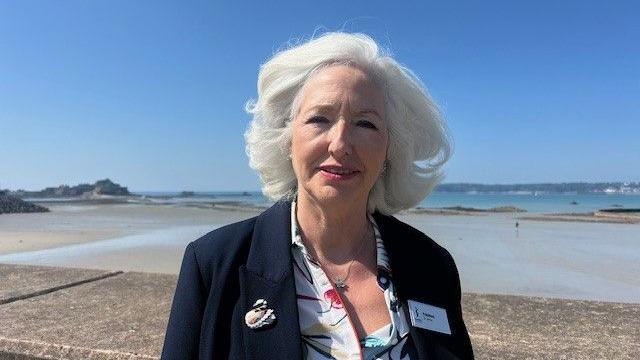 Debbie Taylor is wearing a dark blue blazer and a white shirt with floral patterns. She has white hair and is stood by the sea with Elizabeth castle in the background.