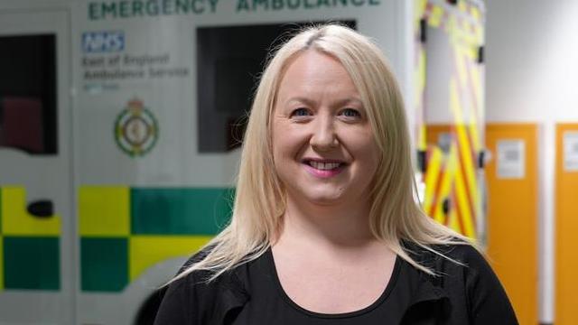 Kerry Gaskin was looking at the camera smiling. She had long blonde hair and was wearing pink lipstick and a black top. She was standing in front of a white ambulance which had a yellow and green chequered pattern in the middle.