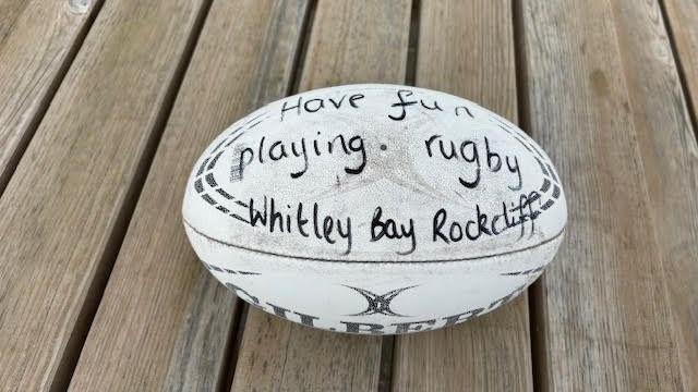 A rugby ball lying on wooden decking. The ball is a white Gilbert ball and appears well worn. A message has been written on in black market pen. It reads, Have fun playing rugby - Whitley Bay Rockcliff.