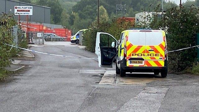 A police van is pictured in an industrial estate, with police cordon tape spanning the width of the road