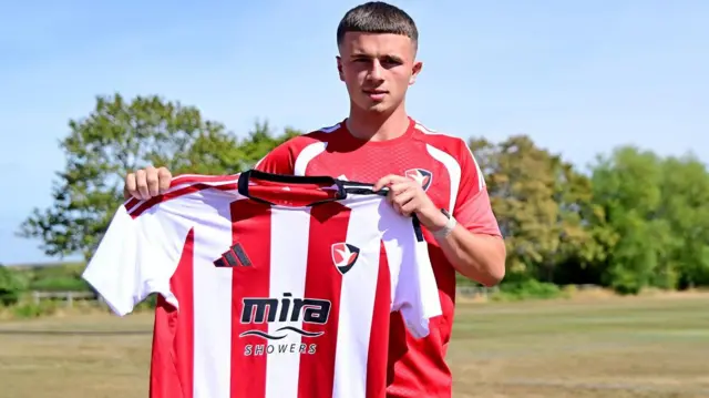 Jake Evans in red Cheltenham Town training top holds up a red and white striped Cheltenham kit with grass and trees behind him.