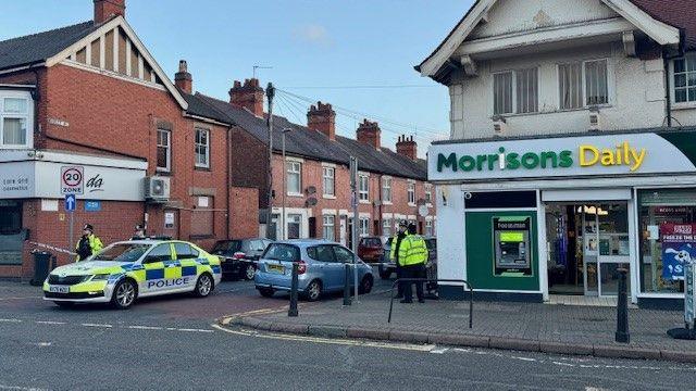Police car parked behind cordon tape at the end of a city street with police stood on the pavement and outside a corner shop