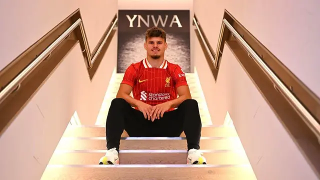 Milos Kerkez sits on a flight of steps with the letters YNWA on the wall at the top, behind his head