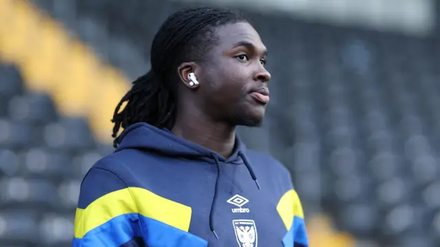 Osman Foyo head and torso shot as he looks thoughtful whilst walking across a pitch with ear buds in. He is wearing a dark blue AFC Wimbledon tracksuit with yellow and light blue chevrons on both sleeves.