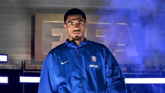 Levi Colwill looks stern-faced wearing a blue Chelsea jacket at an indoor event with blue smoke in the background