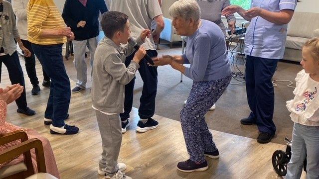 Lucciano wears a grey tracksuit and holds a microphone as he sings. He's joined by residents on their feet, one of whom is dancing next to him as she makes eye contact with him.