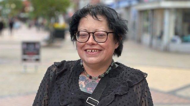 A woman with short black hair and glasses smiles into the camera. She has a necklace made of coloured beds and is wearing a black blouse. A tattoo is also visible on her chest.