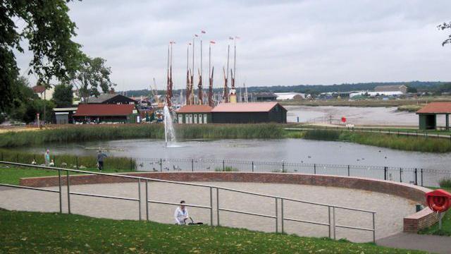 The Maldon amphitheatre in front of a body of water, pedestrians passing by, in the background is an estuary and tall flags