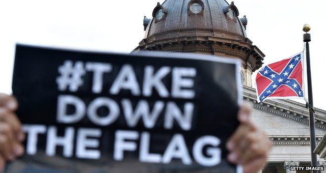 A sign reading "hashtag take down the flag" is held by a protestor with the flag in the background