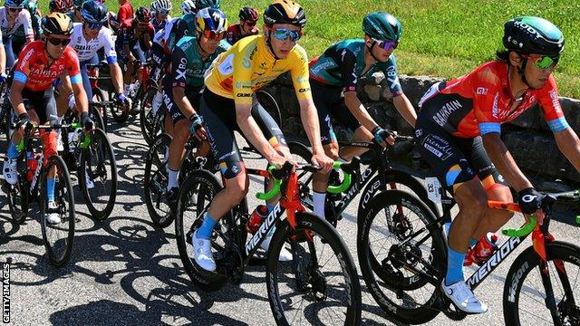 Stephen Williams, in the yellow leader jersey, Yukiya Arashiro of Japan and Team Bahrain Victorious and the peloton compete during the Tour de Suisse 2022