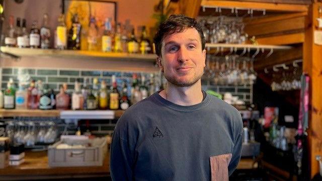 James Ince, owner of Number 8 Cocktail Bar pictured wearing a dark blue shirt. He is stood behind his bar with spirit bottles in the background. 