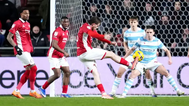Max Bird (centre) stands on one leg with his right leg stretched out to the side after striking the ball towards the goal 