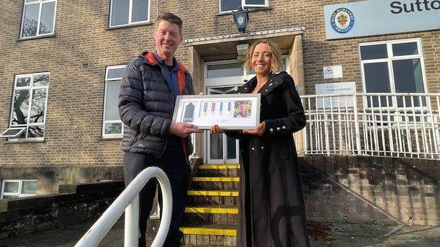 A man and a woman stood outside a police station holding framed police medals.