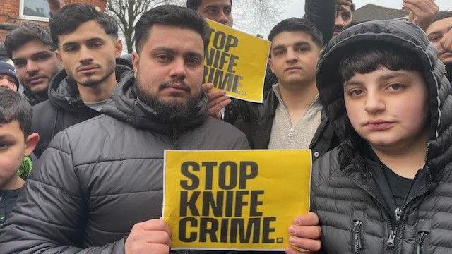 A group of male protesters, mainly wearing dark coats and some holding up yellow placards reading "Stop Knife Crime". 