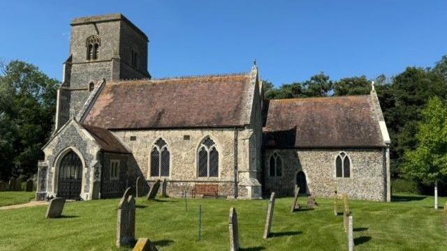 A church with stone coloured walls, red tiled roof, and grey coloured entrance and tower. There are gravestones in the churchyard with green grass.