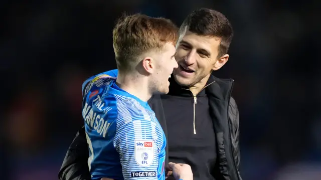A photo of John Mousinho, wearing a black zip-up jumper and a black coat, embracing Zak Swanson in his blue Portsmouth kit after a match in January 2023