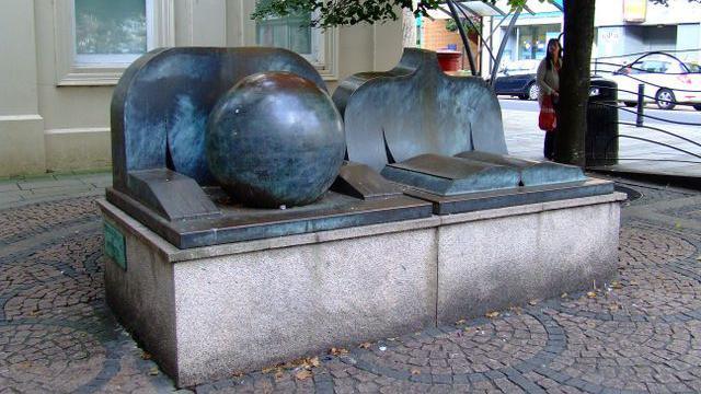 A large sculpture on a street, showing an open book, a large globe and two chests and arms behind them. 