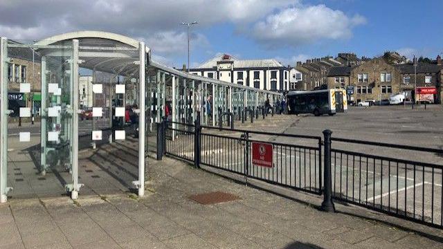 A long bus shelter with a railing in the foreground and a concrete area behind with a bus in the distance. There are buildings in the background.