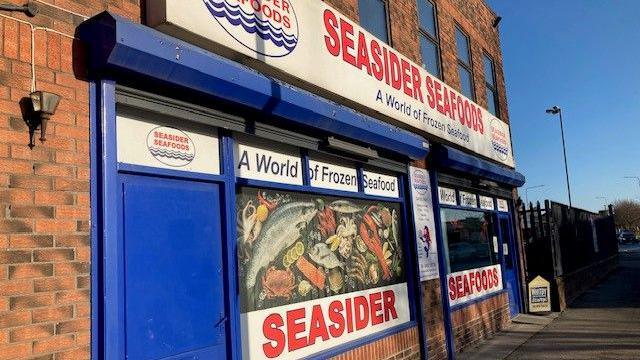 A redbrick building with blue door. There are two large signs in red text, which read: "Seasider Seafoods". A picture of fish and seafood covers one window. Another slogan reads: "A world of frozen seafood".