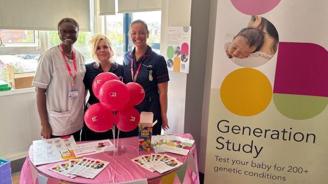 Research Assistant Practitoner Folasade Olufemi-Ajayi and Research Midwives Lucy Rowland and Kirsten Herdman smiling in front of a table with leaflets about the study. There is colourful banner reading Generation Study next to them. Folasade Olufemi-Ajayi has short black hair and glasses. Lucy Rowland's blonde hair is in bun. Kirsten Herdman's brown hair is also in a bun at the back of her head. They are all wearing medical scrubs.