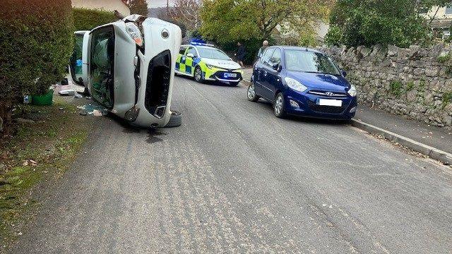 A car lies on its side on a road with police parked behind it. There are people walking in the background and a blue car is parked on the side of the road.