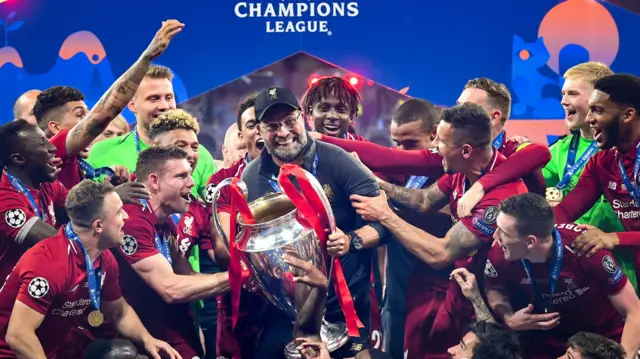 A smiling Jurgen Klopp, wearing a dark cap and top, about to lift the Champions League trophy surrounded by celebrating Liverpool players in 2019