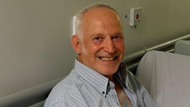 Patient Louis Bella in a hospital corridor. He is smiling and his shirt is unbuttoned and he has a small plaster on his chest. 