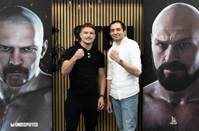 Two men - one in a black t-shirt and one in a white button-up shirt and jeans, stand in front of a wood-panelled wall pulling a boxing pose with a cocked fist.