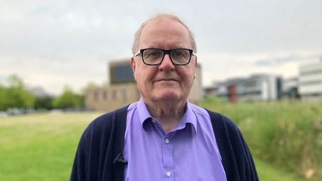 Prof Ian Barnes wearing a lilac shirt and dark cardigan and dark framed glasses stands in front of buildings at the University of Lincoln.