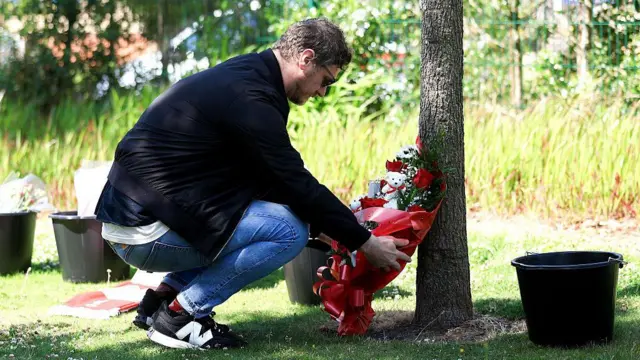A man in a black jacket lays a bouquet of flowers next to a tree trunk