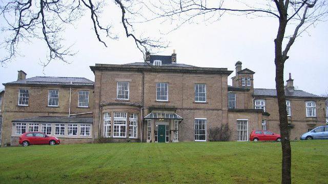 Shotley Park is a large two-storey house with big windows and a green entrance door. There are three parked cars in front of it and grassland.
