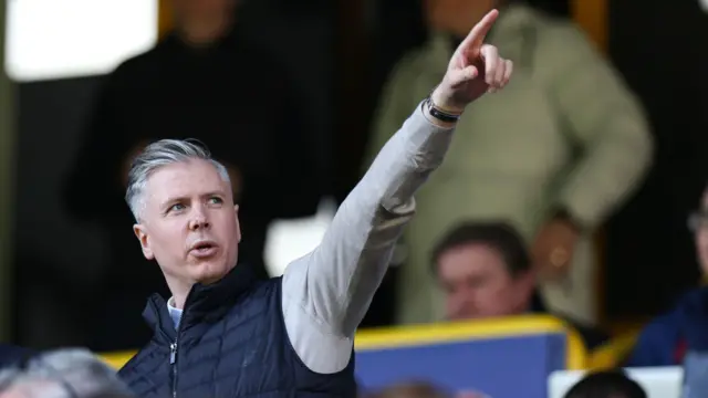 A photo of Lee Bromby, wearing a black gilet over the top of a grey jumper, pointing up to the sky from a football stand.