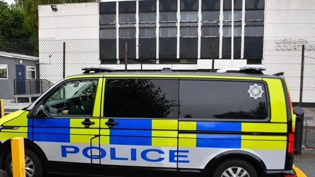 A police van is parked in front of Lisburn Court House. The windows are heavily tinted. The car is yellow and blue with POLICE in large lettering. The court house is painted white.