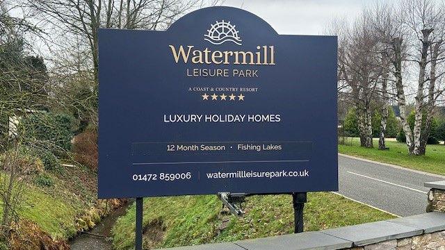 A blue sign at the entrance to Watermill Leisure Park, advertising luxury holiday homes, with trees in the background. 