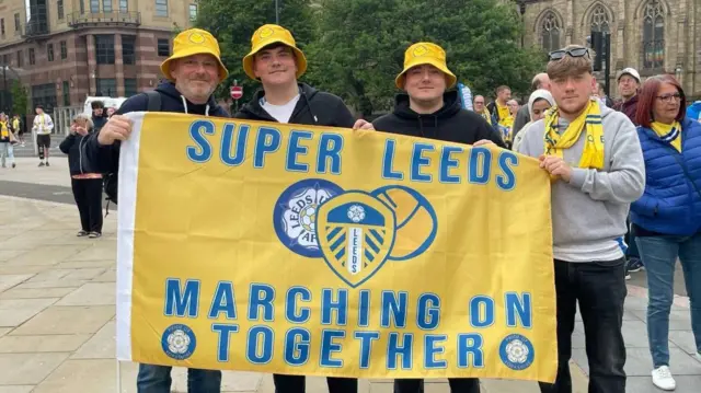 A father poses for a picture with his three sons, all holding a Leeds United flag that reads: 'Super Leeds - Marching On Together'