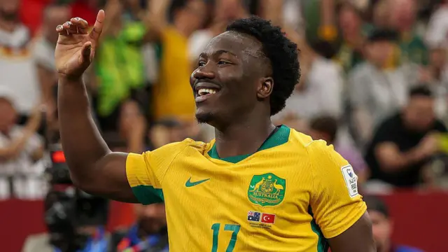A smiling Nestory Irankunda wearing a yellow Australia playing shirt with the number 17 on it celebrates his goal against Turkey with his right arm raised and pointing his index finger to the sky
