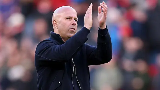 Arne Slot, wearing a dark jacket, applauds towards fans after a match. The crowd is out of focus in the background.