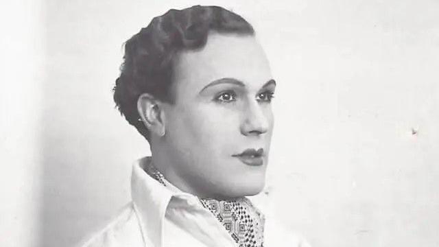 A black and white photo of a young man in profile looking to the right. He has slicked back dark curly hair and is wearing lipstick and eye make up. He's dressed in a white shirt and is wearing a cravat with a geometric pattern
