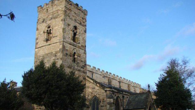The outside of St Cuthbert's which is a stone church with a bell tower at the front. Trees surround the building.