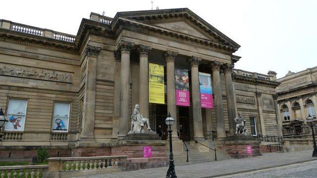 The Georgian facade of the Walker Art Gallery looms over the centre of Liverpool. Street lights, statues and ornate stone fences can be seen.