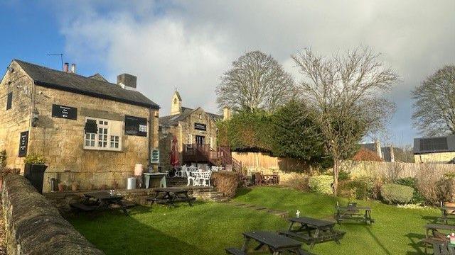 A pub garden, featuring wooden picnic tables, trees and plants