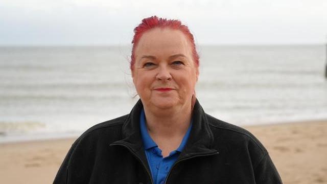 Pam Hallesy has red hair and is wearing a black fleece with the RSPB logo on it. She stands on a beach with the sea behind her.
