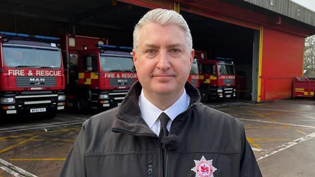 Devon and Somerset Fire Service Gavin Ellis wearing a shirt, tie and a fire service coat. He is stood in front of a fire station with four fire engines inside.