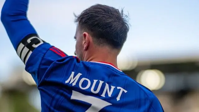 An image of the back of Mason Mount with the stands out of focus in the background. He is wearing the captain's armband and holding his arm in the air.