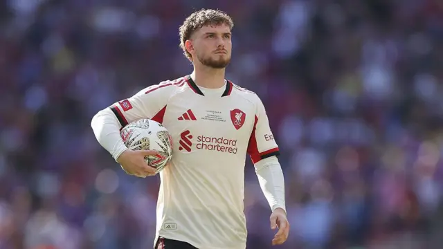 Harvey Elliot wearing Liverpool's away kit and holding a football