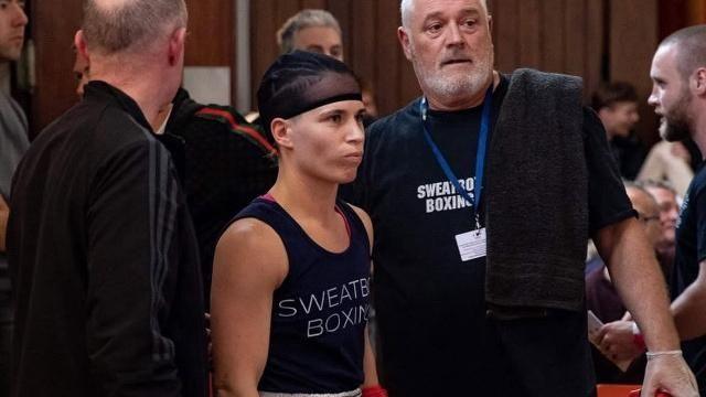 A female boxer wearing a cap and blue shirt has her cornerman by her side with a black towel over his shoulder. They are both in a crowded room.