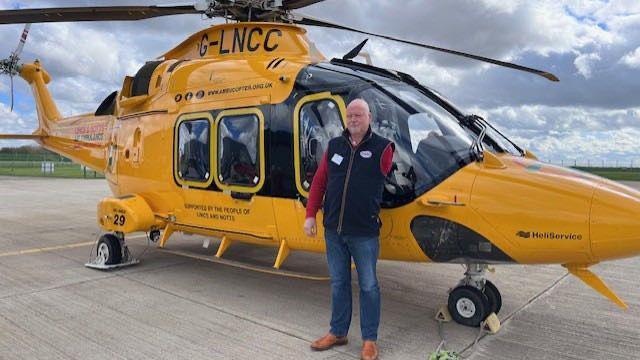 A bald man stands outside next to a yellow helicopter. He has one arm missing on his left side