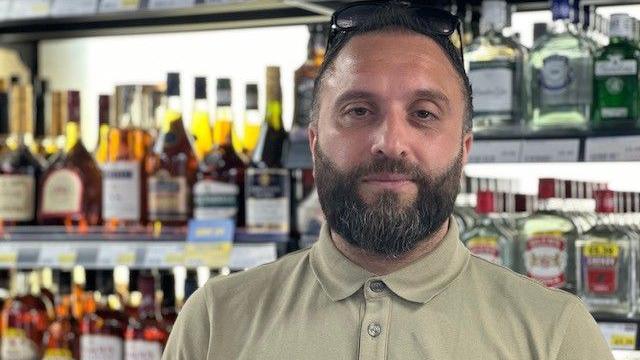 A man with a dark beard and hair wears a stone-coloured polo top. He is standing in front of bottles of alcohol